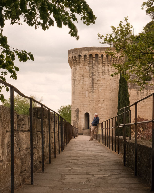 A lone man leans against the rail of a stone pathway beside a tall, circular tower in Avignon, framed by trees and historic ramparts on a cloudy afternoon.