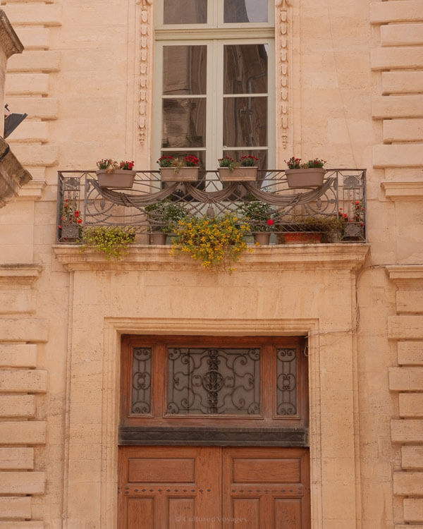 Image of a charming upper-floor balcony in Avignon, decorated with terracotta pots filled with blooming red and yellow flowers, positioned above an ornate wooden door with wrought iron detailing.