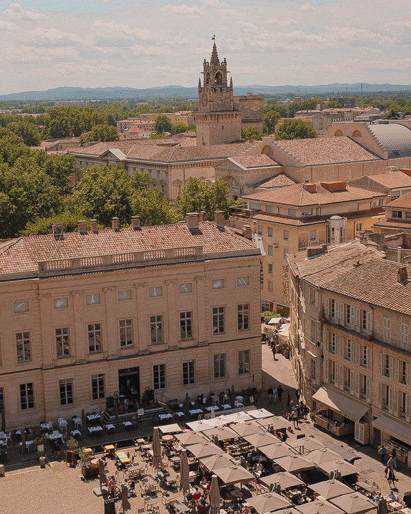 Panoramic view over Avignon’s rooftops and central square, showing the historic bell tower of the Hôtel de Ville rising above surrounding stone buildings and umbrella-shaded outdoor cafés below.