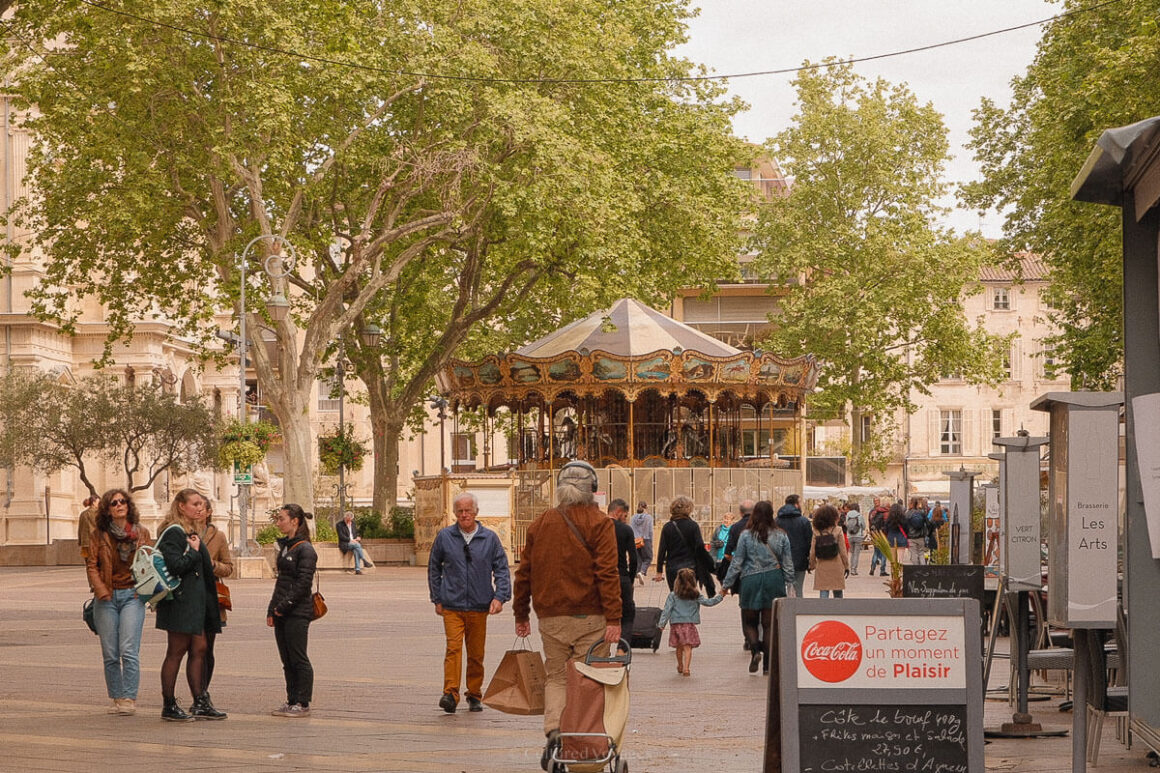 A classic carousel with ornate decorations sits in the centre of a busy plaza, surrounded by leafy trees and people strolling, shopping, or chatting.
