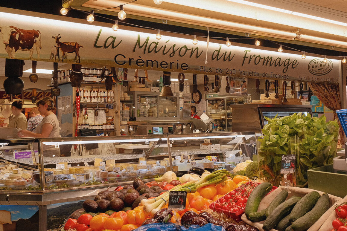 Inside a bustling French market stall labelled “La Maison du Fromage”, showcasing a variety of cheeses behind glass and a colourful array of fresh produce in the foreground. A delightful snapshot from one day in Avignon.