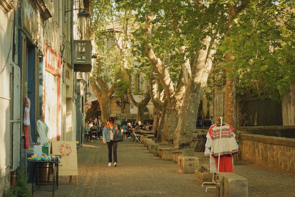 A cobbled pedestrian street shaded by large plane trees, with cafés and shops lining the left side and embroidered garments displayed outside; a woman walks past while others sit at outdoor tables.