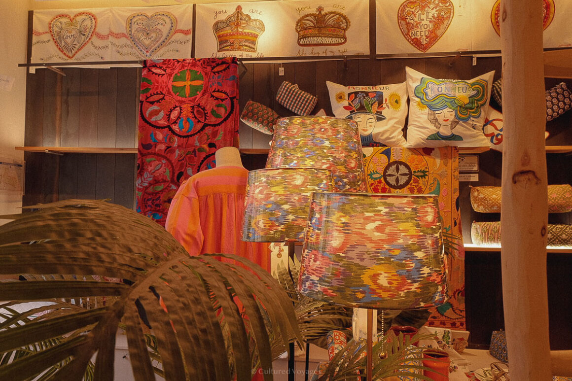Interior of a boutique shop with vibrant, patterned lampshades and textiles, including illustrated pillows, embroidered hearts and crowns, and a mannequin dressed in a colourful robe.