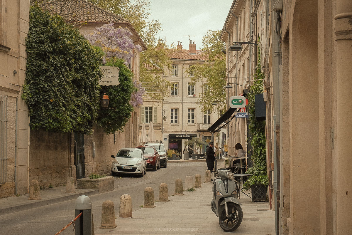 A quiet street in Avignon with cars parked along the kerb, wisteria draped from a building, and a sign for Hôtel d’Europe hanging beside a lamppost wrapped in ivy.