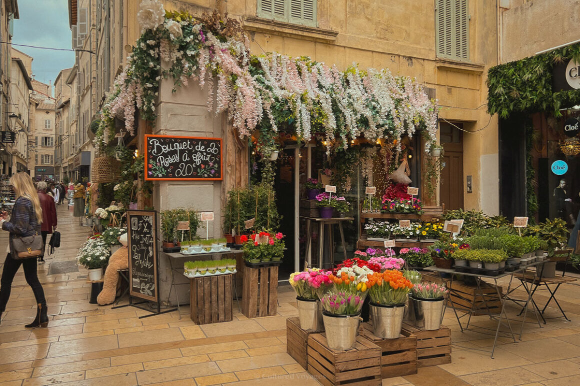 An abundant florist shop on a stone-paved street, with buckets of colourful flowers, cascading blossoms overhead, and signs written in French advertising bouquets.