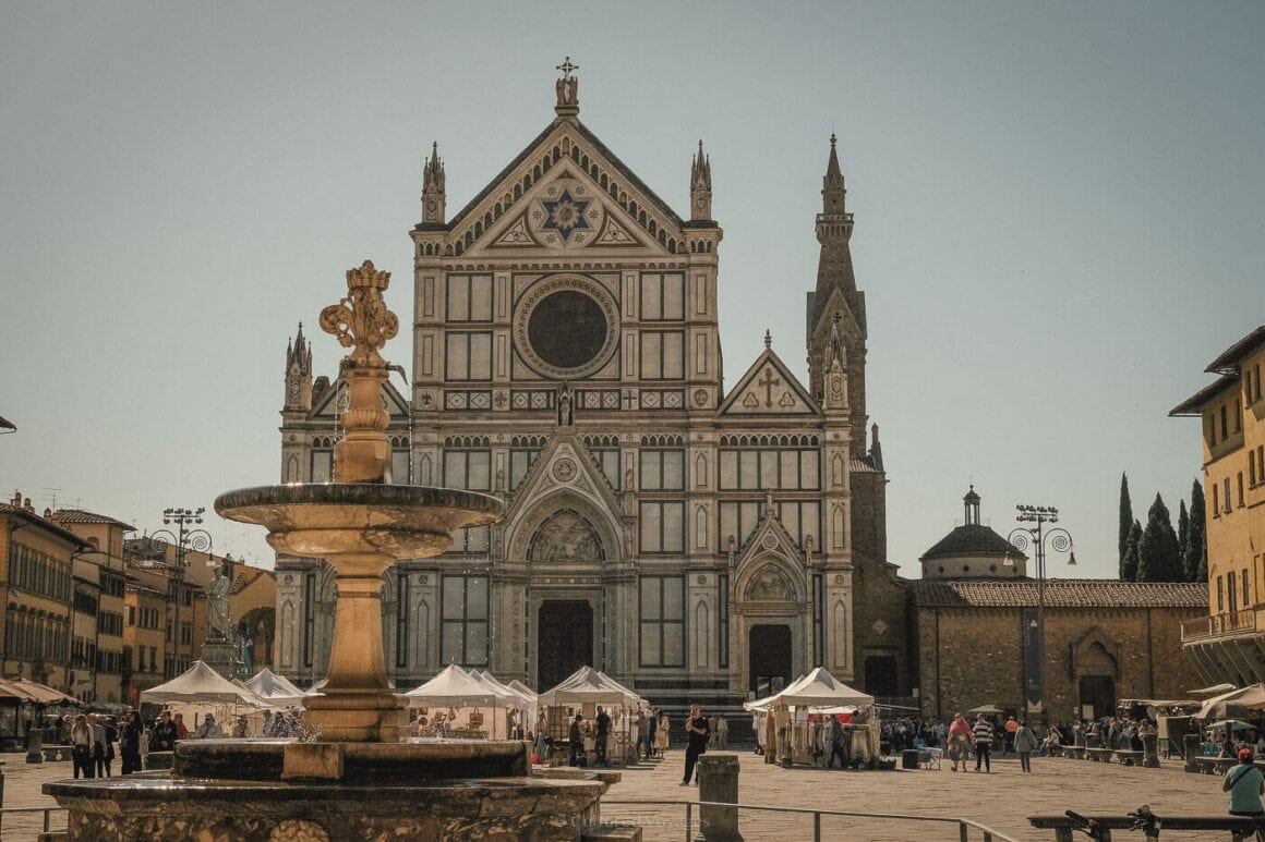 Historic Santa Croce Basilica and the stone fountain in its lively piazza, surrounded by market tents and classic Florentine architecture.