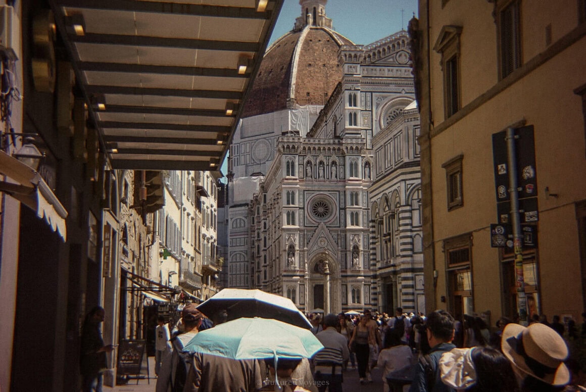 Crowds with umbrellas walk along a narrow Florence street with the Florence Cathedral dome towering at the end, showing how busy Florence has become in May.