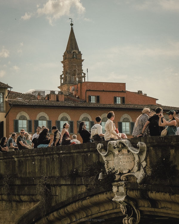 Group of tourists relaxing on the stone railing of Ponte Santa Trinita as the bell tower rises in the backgroundโcapturing spontaneous charm during the best time to visit Florence.