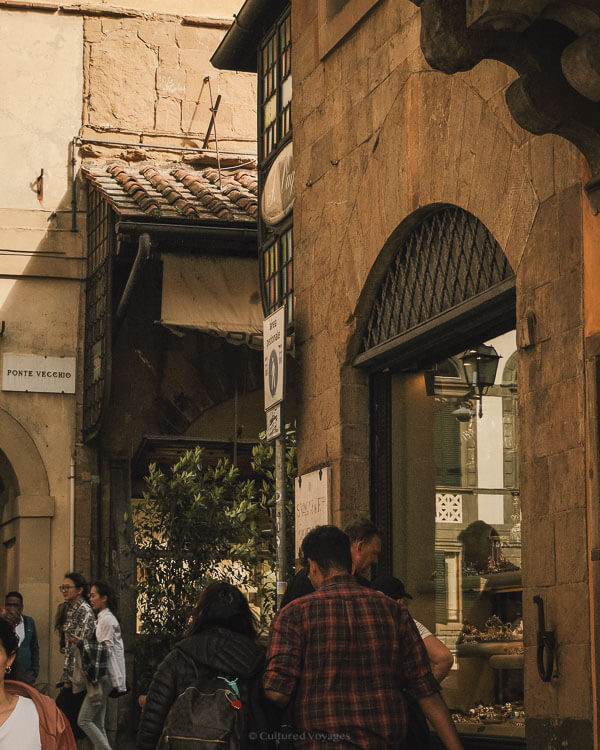 Locals and tourists stroll past jewelry shops along the Ponte Vecchio, under soft afternoon light