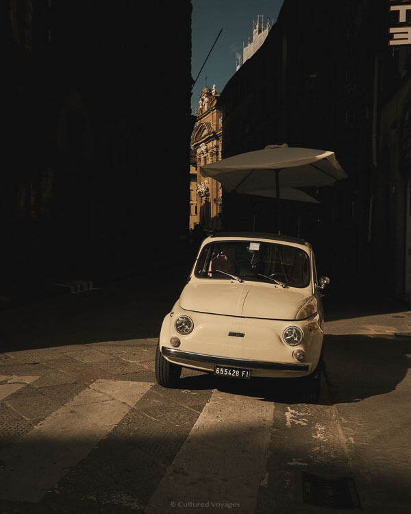 A classic white Fiat 500 parked under a street umbrella in a shadowed Florentine alleyway, radiating nostalgic charm.