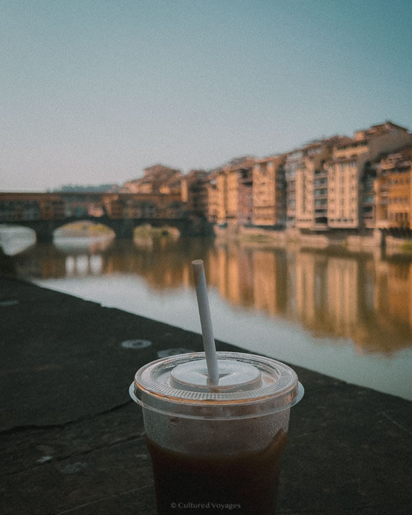 A plastic cup of iced coffee set against the backdrop of Ponte Vecchio and the Arno River, perfect for enjoying the best time to visit Florence.