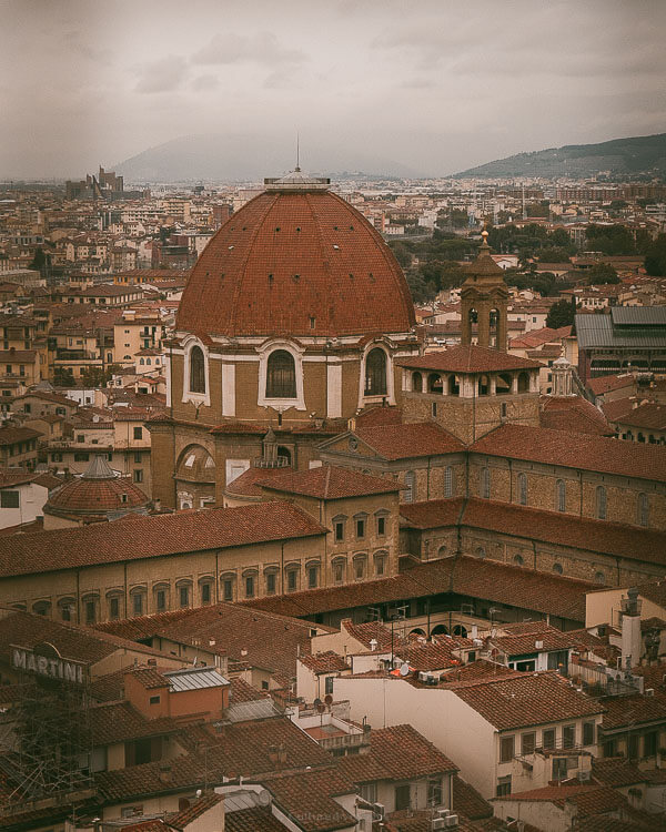 Aerial view of the red dome of San Lorenzo church surrounded by Florenceโs rustic rooftops under a moody sky.