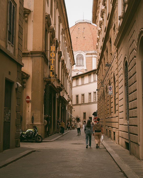 Pedestrians walk through a quiet Florentine alleyway with a historic dome visible in the background.