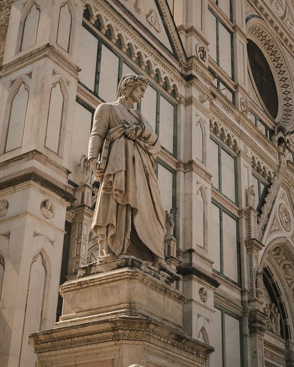 Marble statue of Dante Alighieri standing in front of the richly decorated faรงade of Santa Croce Basilica in Florence.
