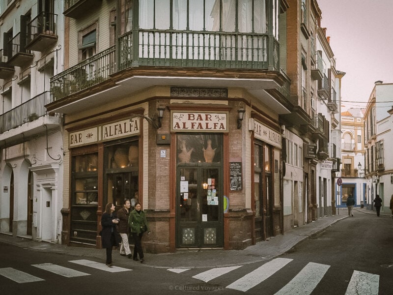 Locals walking past Bar Alfalfa, a corner tapas bar in Seville's historic centre, with old-fashioned signage and warm lighting.