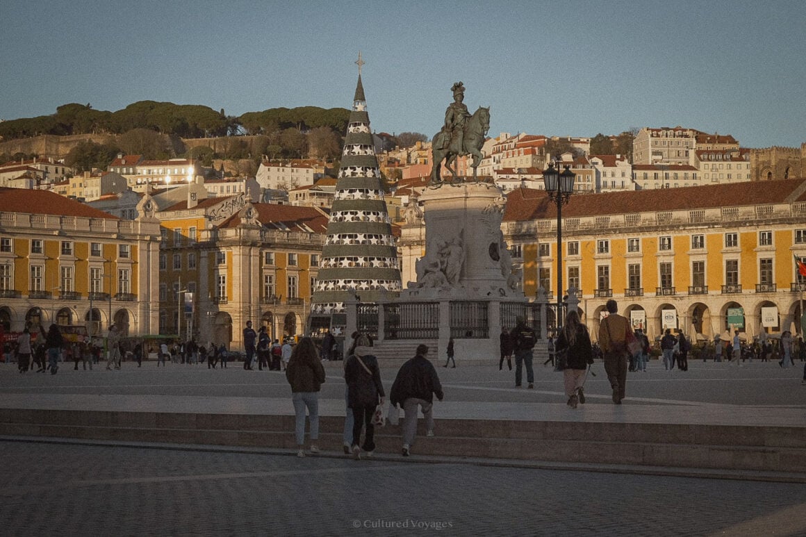 People wander through Lisbon’s Praça do Comércio, where a striking Christmas tree and statue stand against a backdrop of golden-hued buildings under soft winter sunlight.