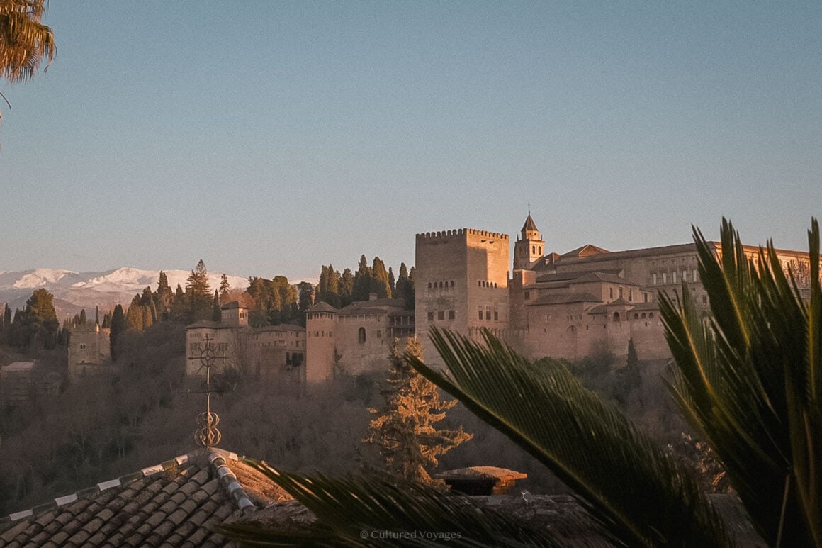 The Alhambra fortress in Granada glows in the soft evening light, framed by palm fronds and snow-capped mountains — a must-see highlight among the best European cities to visit in winter.