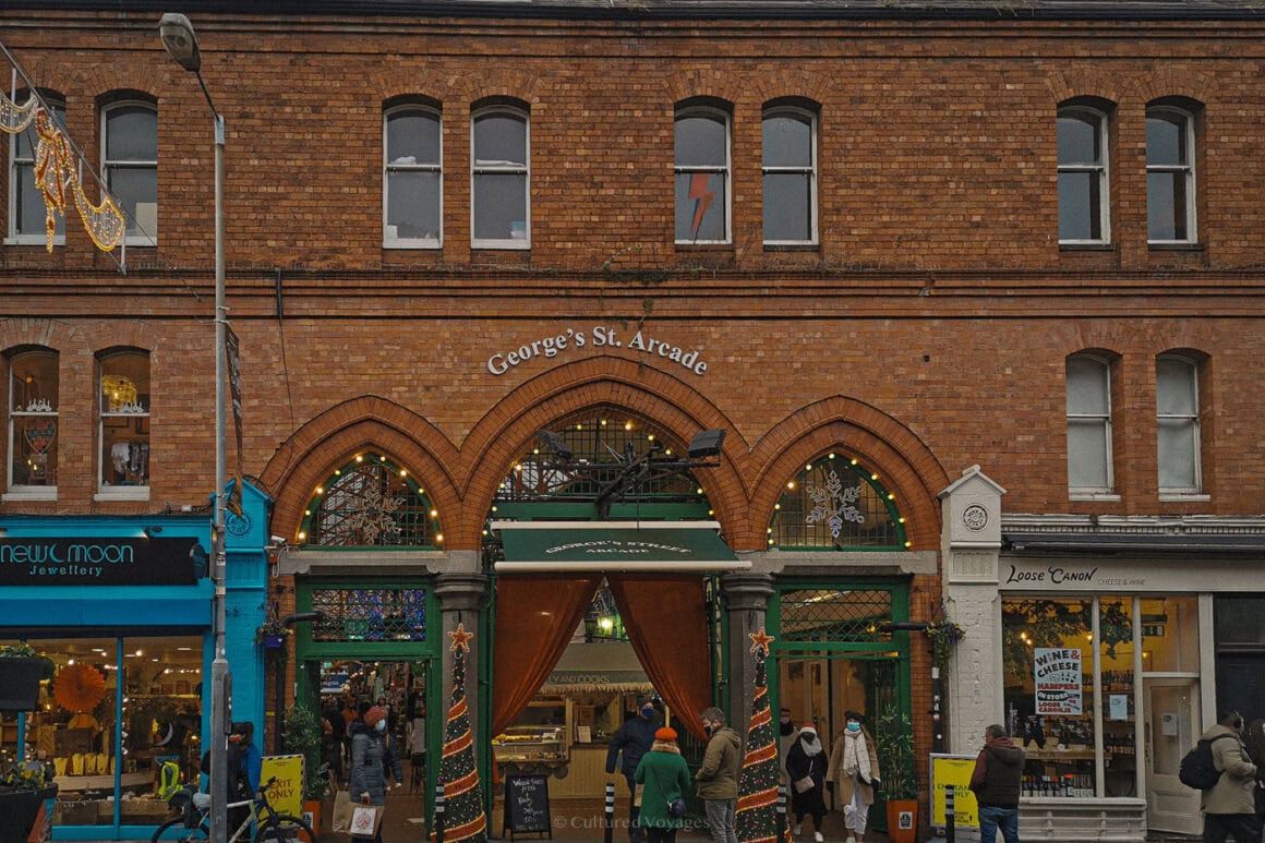 Shoppers pass beneath the festively decorated entrance to George’s Street Arcade in Dublin, a historic indoor market filled with independent shops and cafés during the winter season.