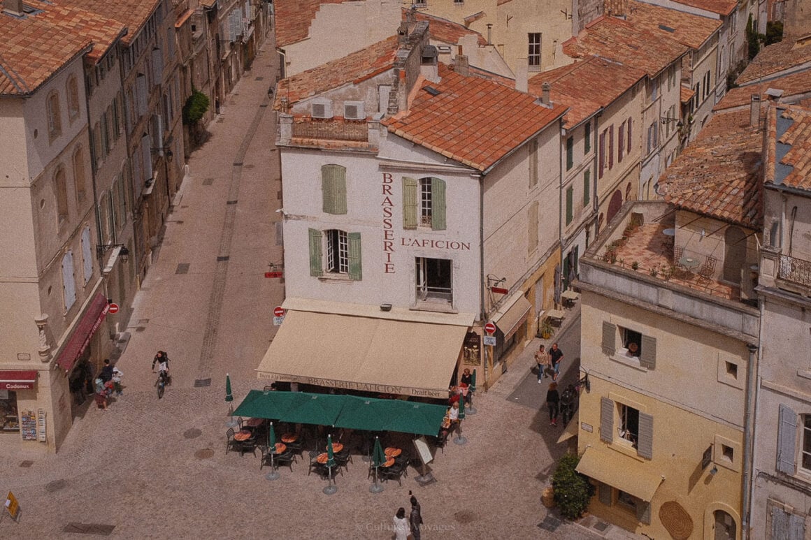A charming aerial view of a quiet street in Arles, France, with a traditional brasserie and café tables beneath green parasols.