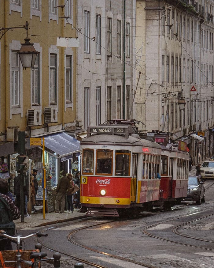 A classic yellow tram winds its way through the historic neighbourhoods of Lisbon, passing locals and shopfronts along cobbled streets.