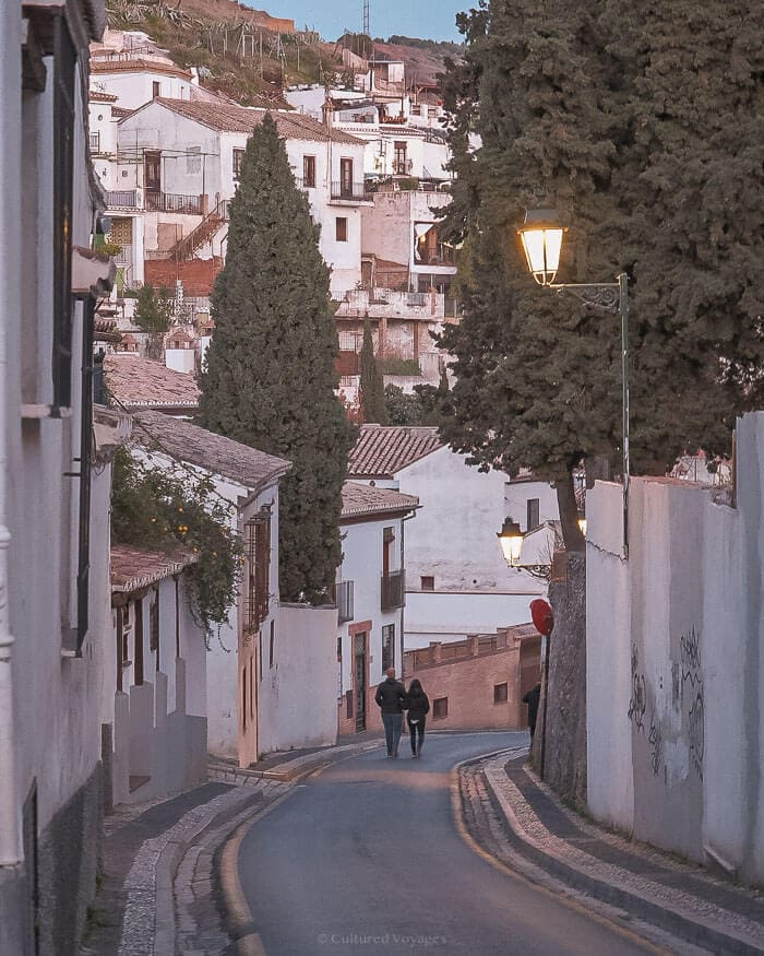 A couple walks down a quiet, winding street lined with traditional whitewashed houses and cypress trees in Granada, Spain.