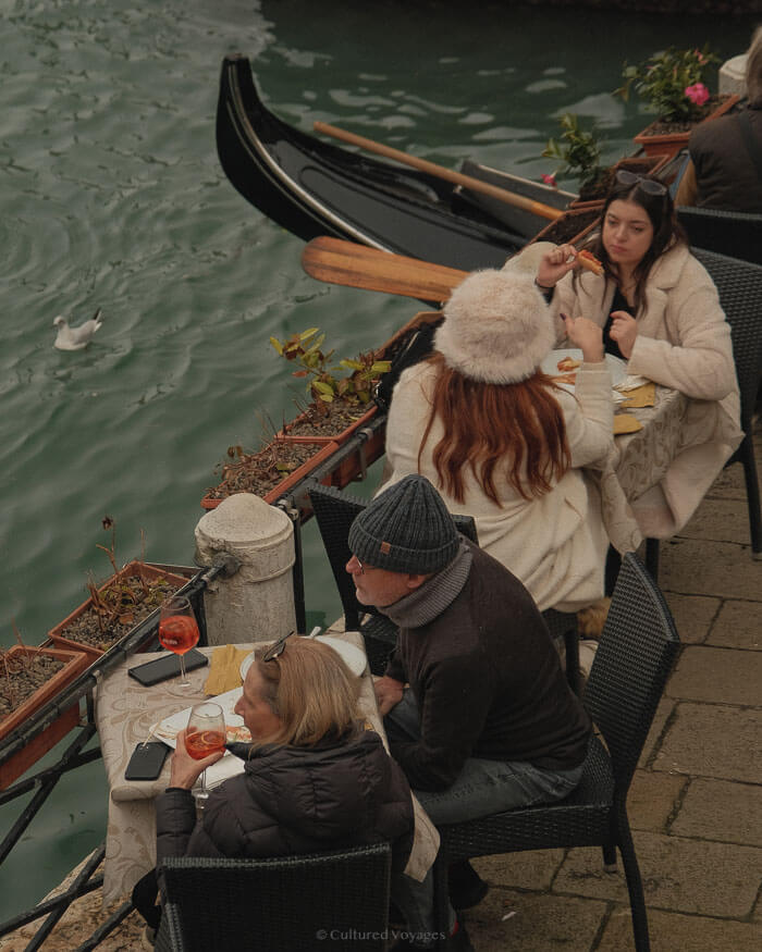 People enjoying lunch by the canal with gondolas passing by in Venice, Italy, capturing the city’s relaxed winter vibe away from the usual crowds.