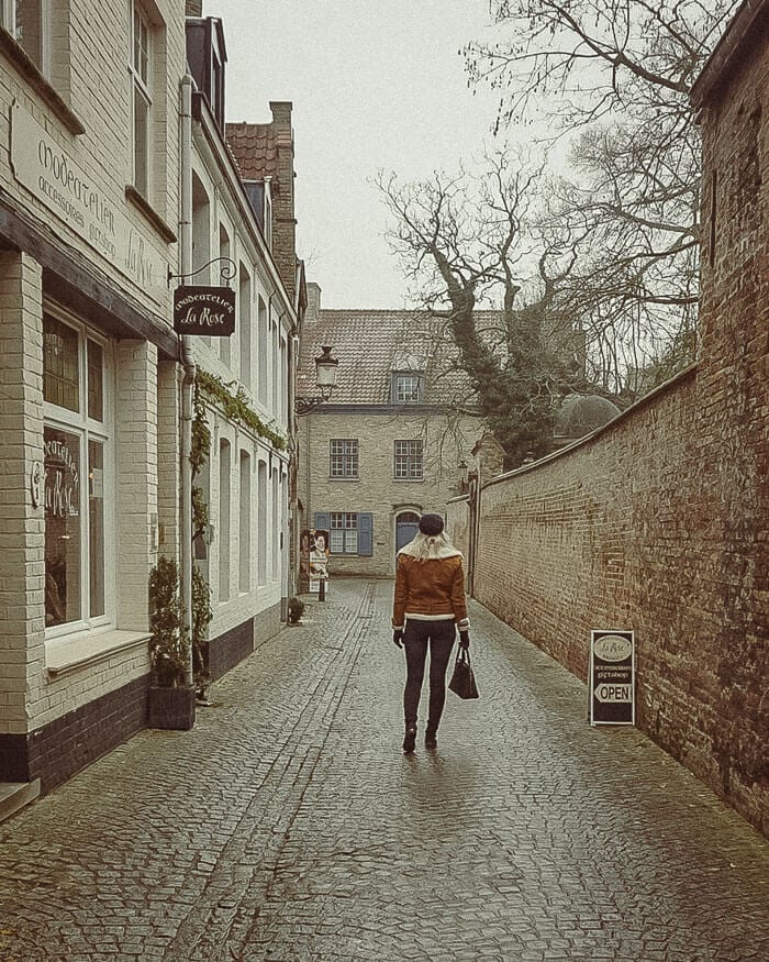 A lone figure walking down a quiet, cobbled lane lined with charming shops in Bruges, Belgium, highlighting the city’s enchanting winter appeal.