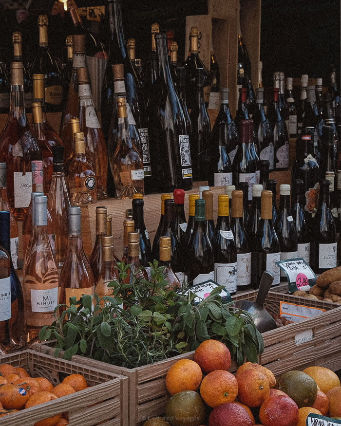 Bottles of wine, citrus fruits, and herbs on display at a lively outdoor market stall in southern France.
