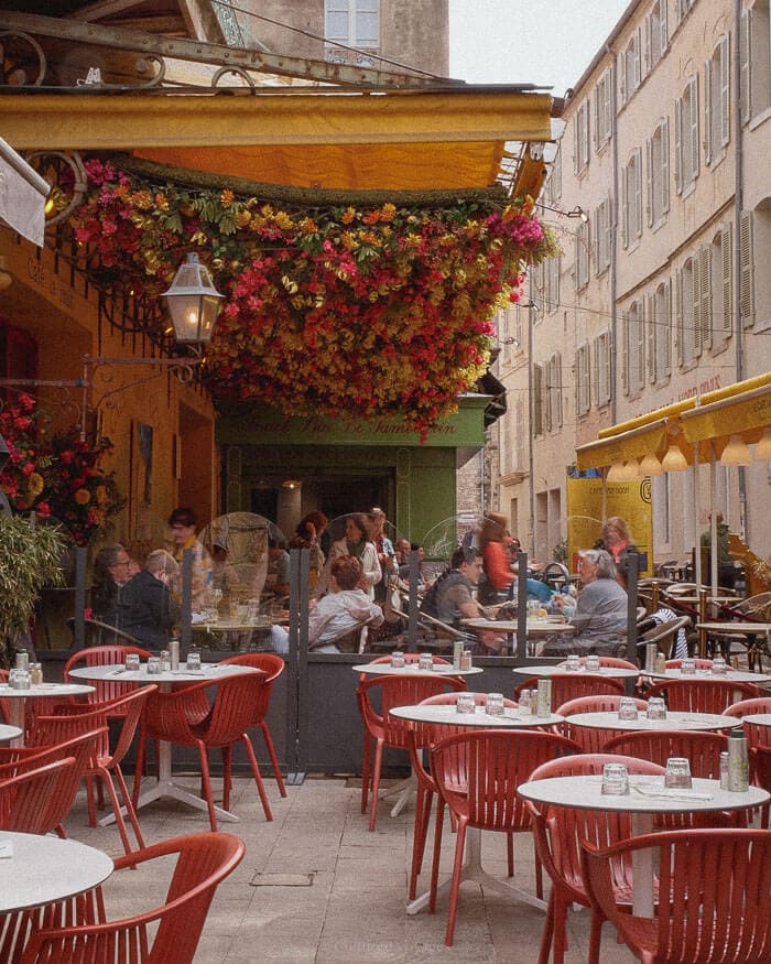 Locals and visitors enjoying lunch at a bustling outdoor café shaded by colourful hanging flowers in a charming French town.