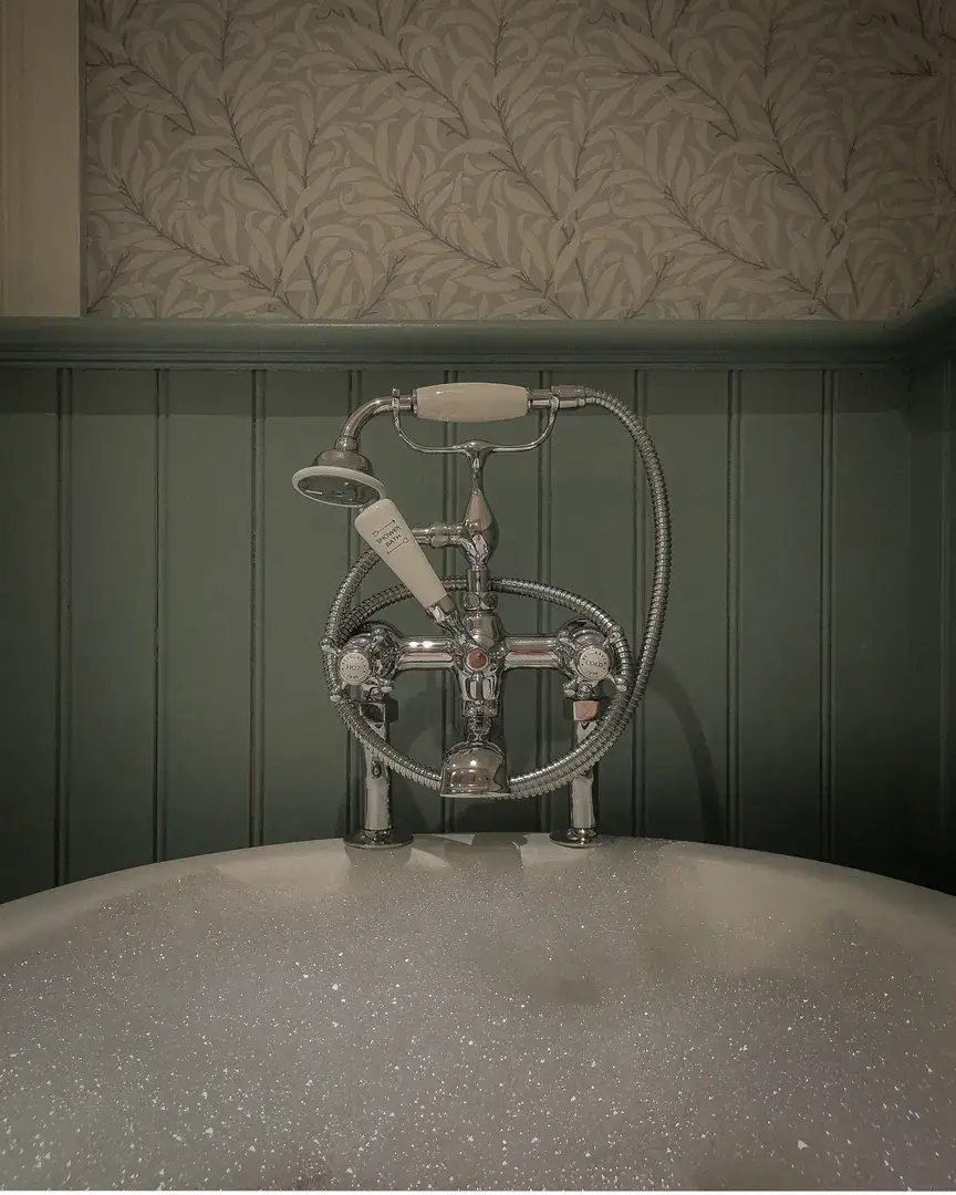 Chrome bath taps above a bubble bath against green panelling and leaf wallpaper
