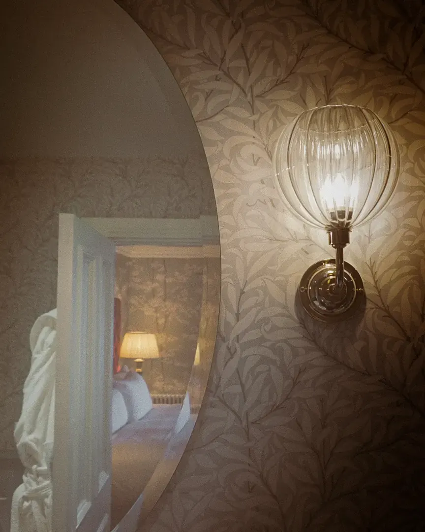 Glass wall sconce beside an oval mirror reflecting the bedroom