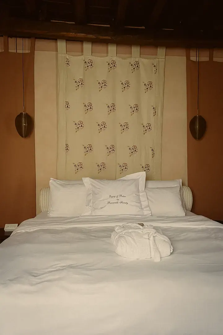 A double bed beneath a printed hanging, with bronze pendant lamps either side and a pillow embroidered with the Romanelli family name