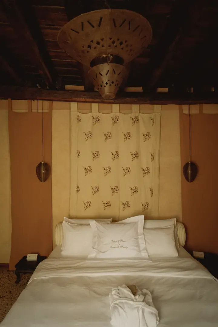 The bed beneath a pierced brass lantern and dark beams, with a printed hanging and pendant lamps either side