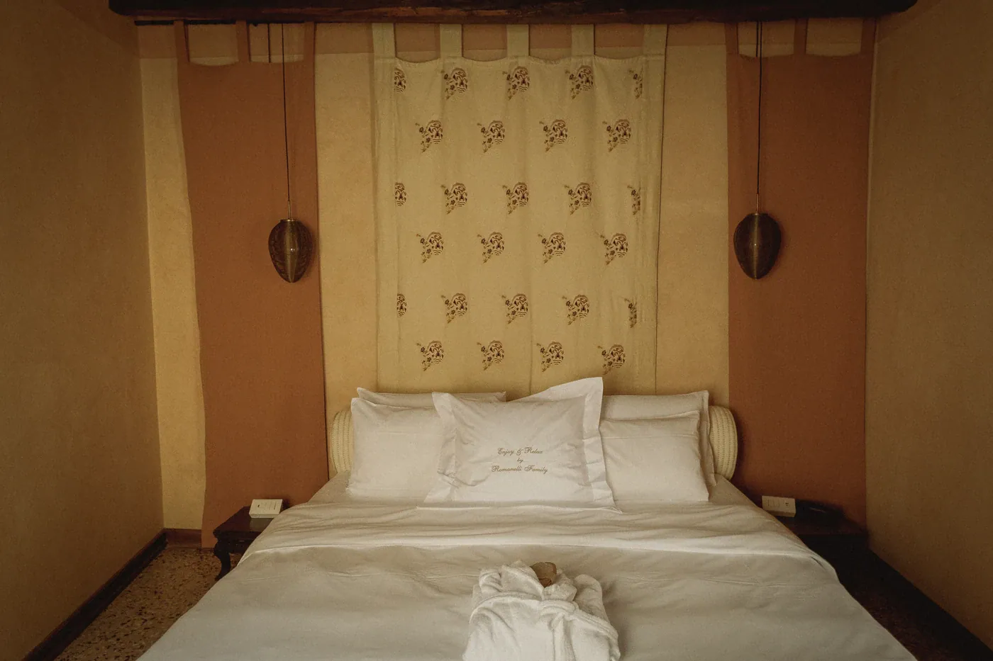 A double room at Novecento, its bed set against terracotta and cream walls beneath a printed hanging, with pendant lamps either side