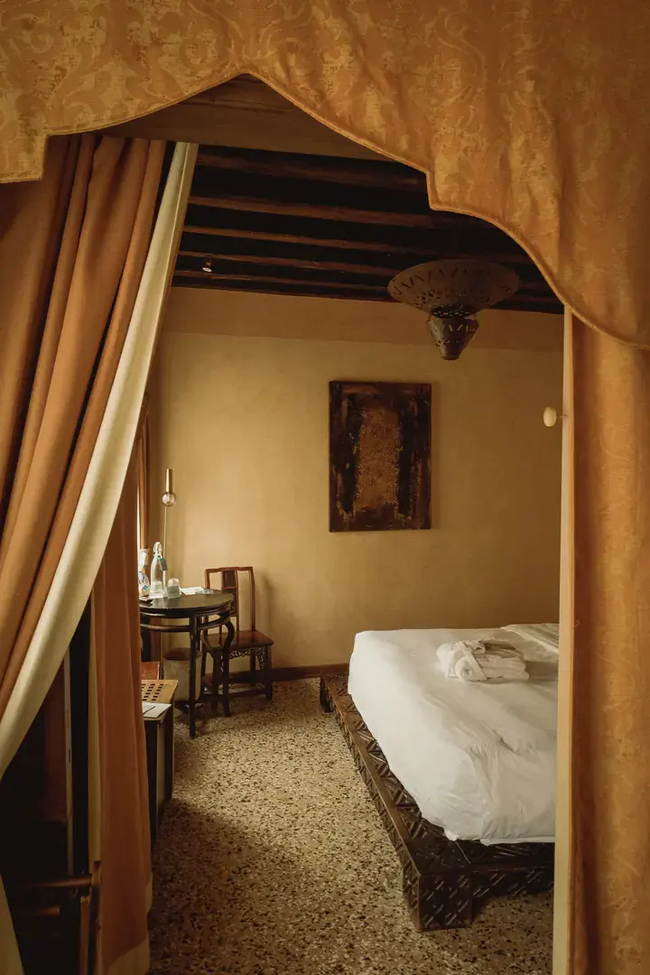 The room seen through a draped archway, with a dark abstract painting, a small round table, a wooden chair and the end of the bed